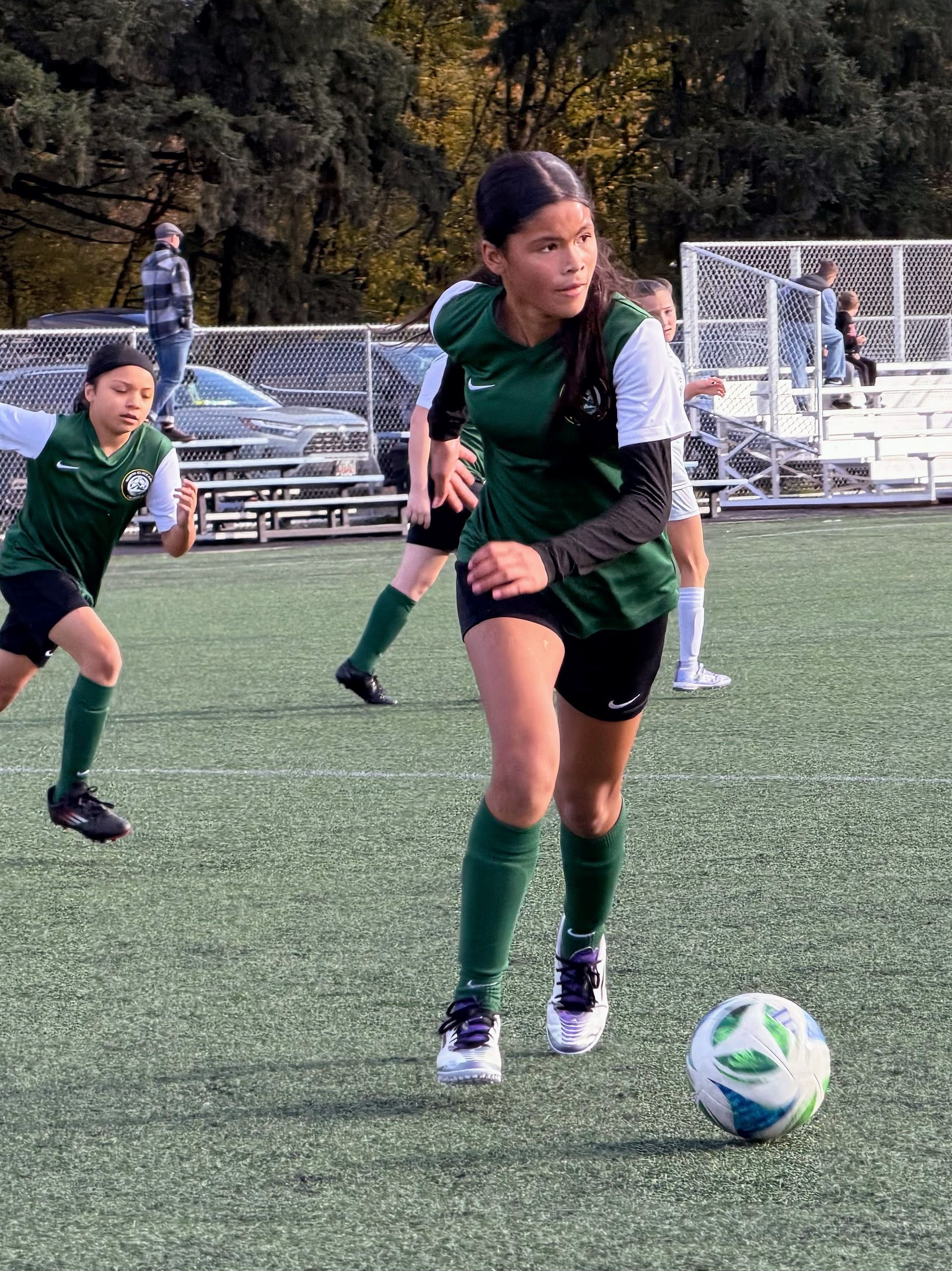 Soccer player in green jersey dribbles a ball on a green field. Others play nearby.