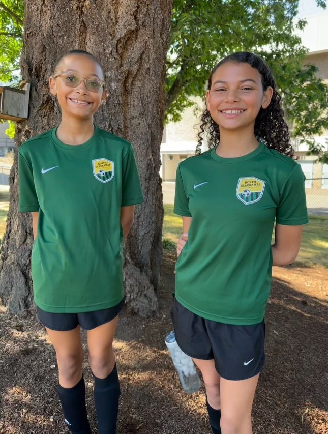 Two people in green soccer jerseys and black shorts stand outside by a tree.