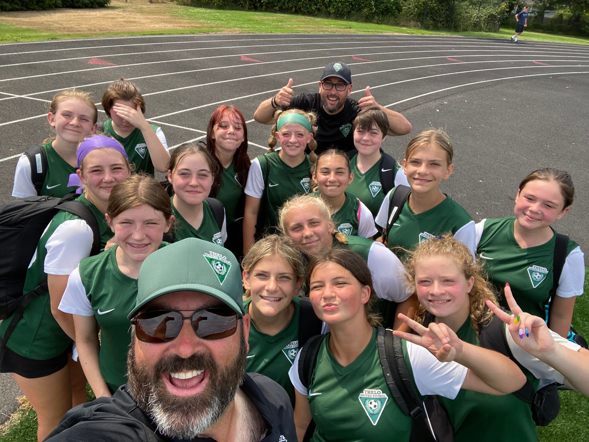 Soccer team in green and white jerseys poses on a track with their coach; some make hand gestures.