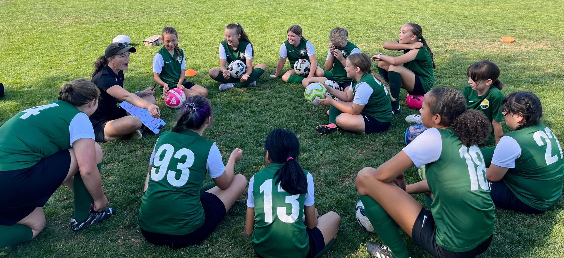 Soccer players in green jerseys sitting in a circle on a grassy field, with the coach giving instructions.