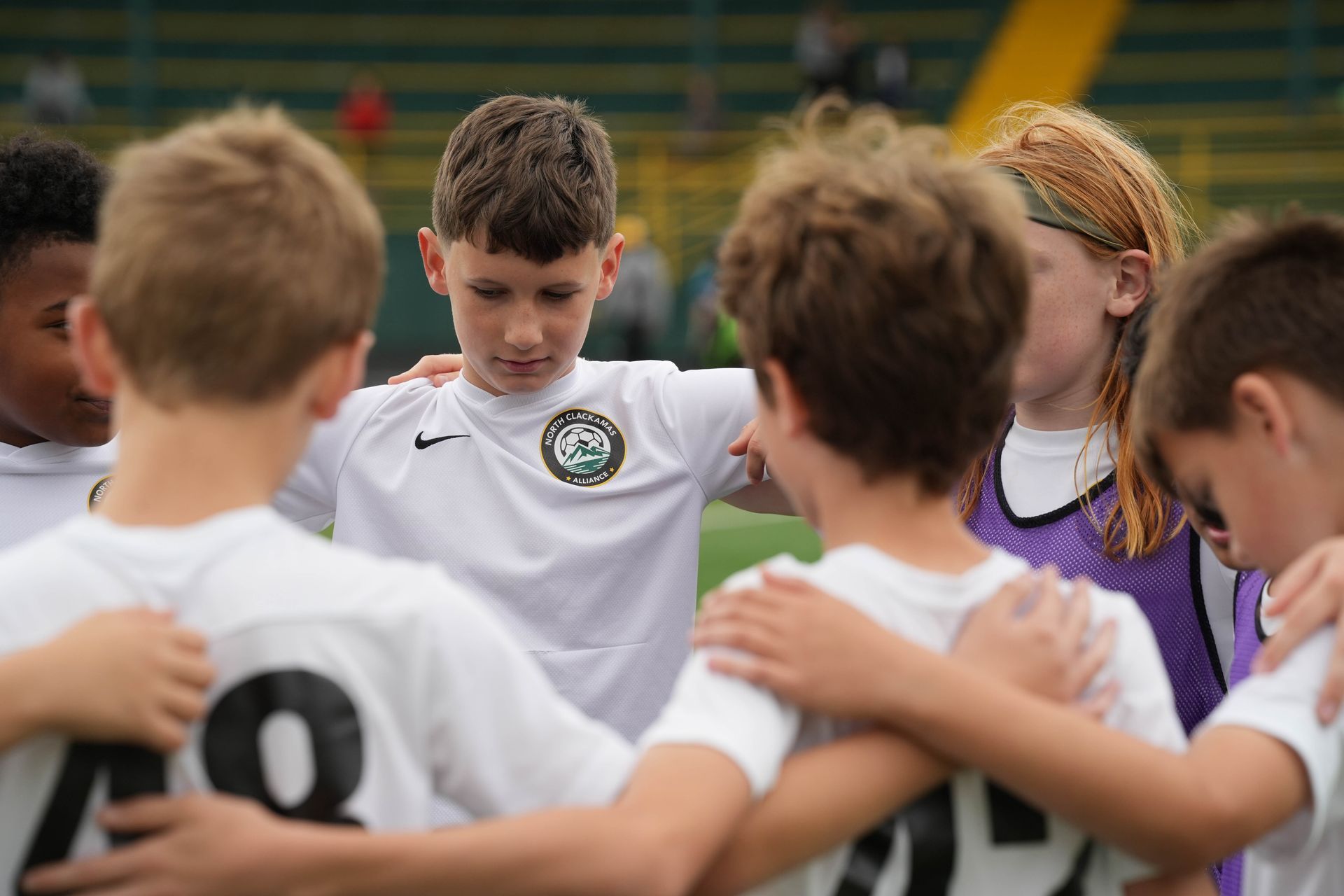 Soccer team huddled together, arms around each other, on a green field. Some wearing white jerseys, looking down.