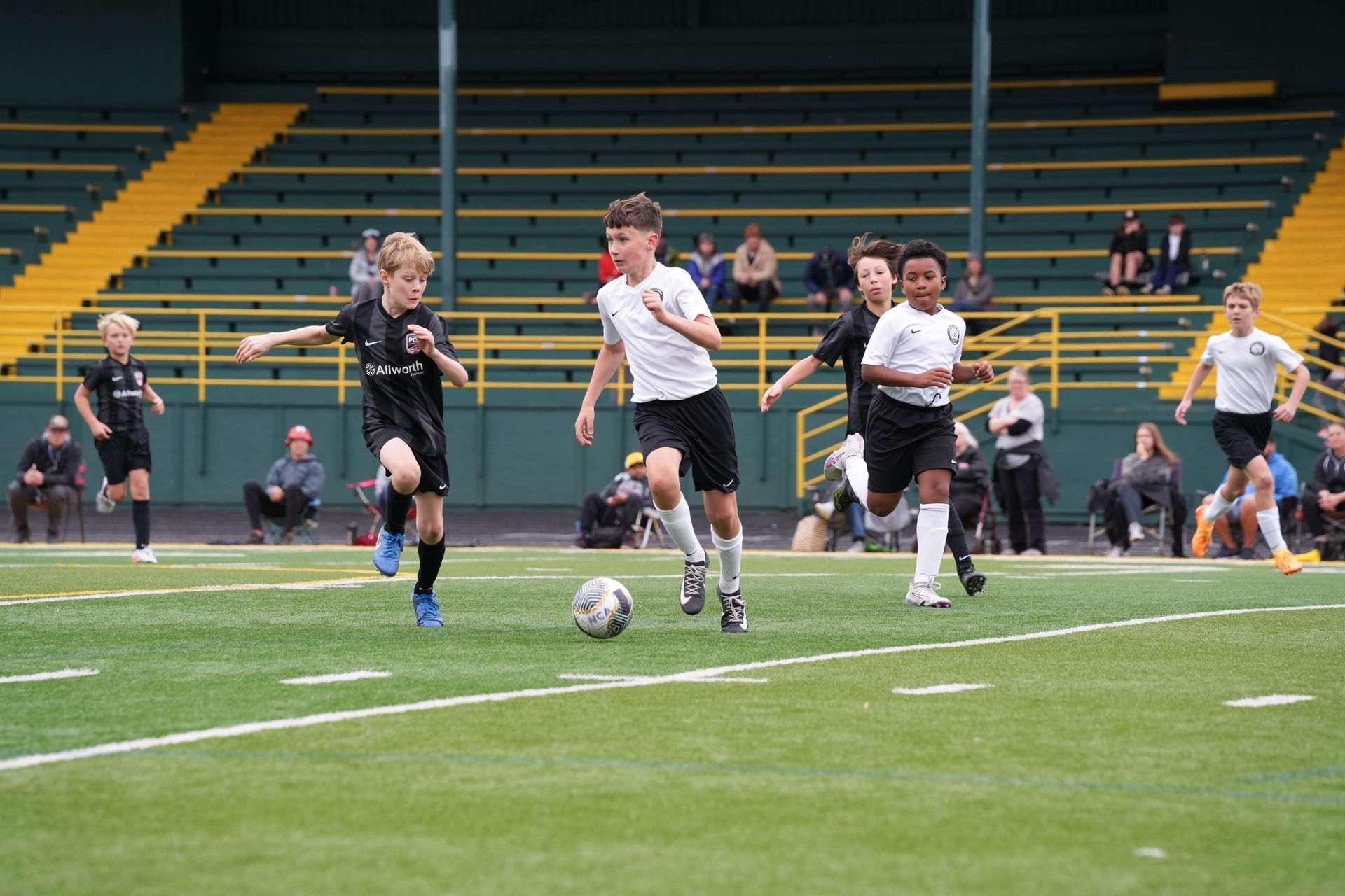 Boys playing soccer on a green field in front of stadium seating; one with the ball, others running.