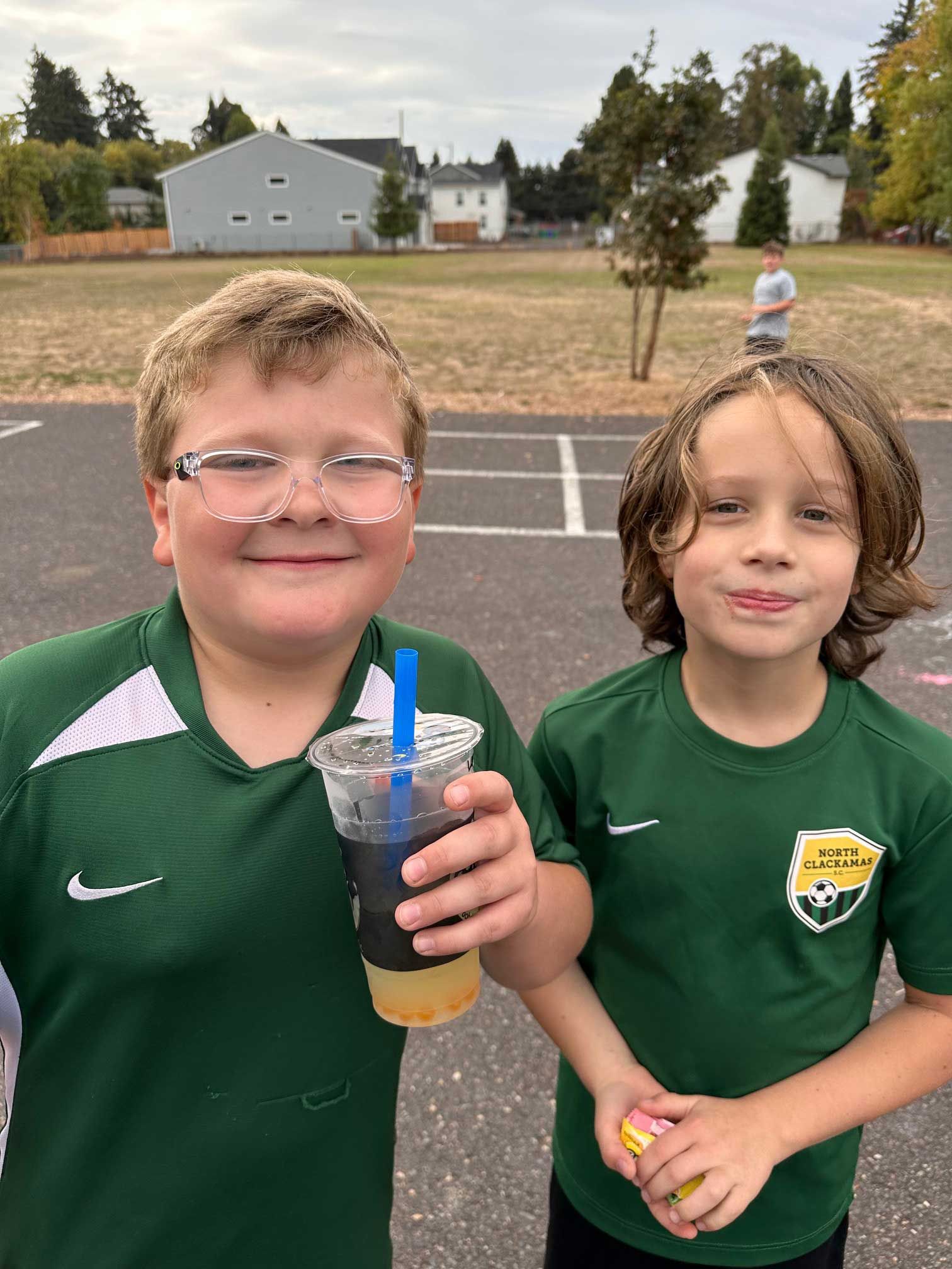 Two boys in green soccer shirts holding a drink and a ball, smiling on a playground.