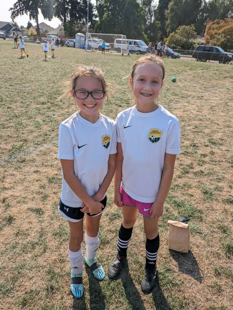 Two girls in soccer uniforms smile on a sunny field.