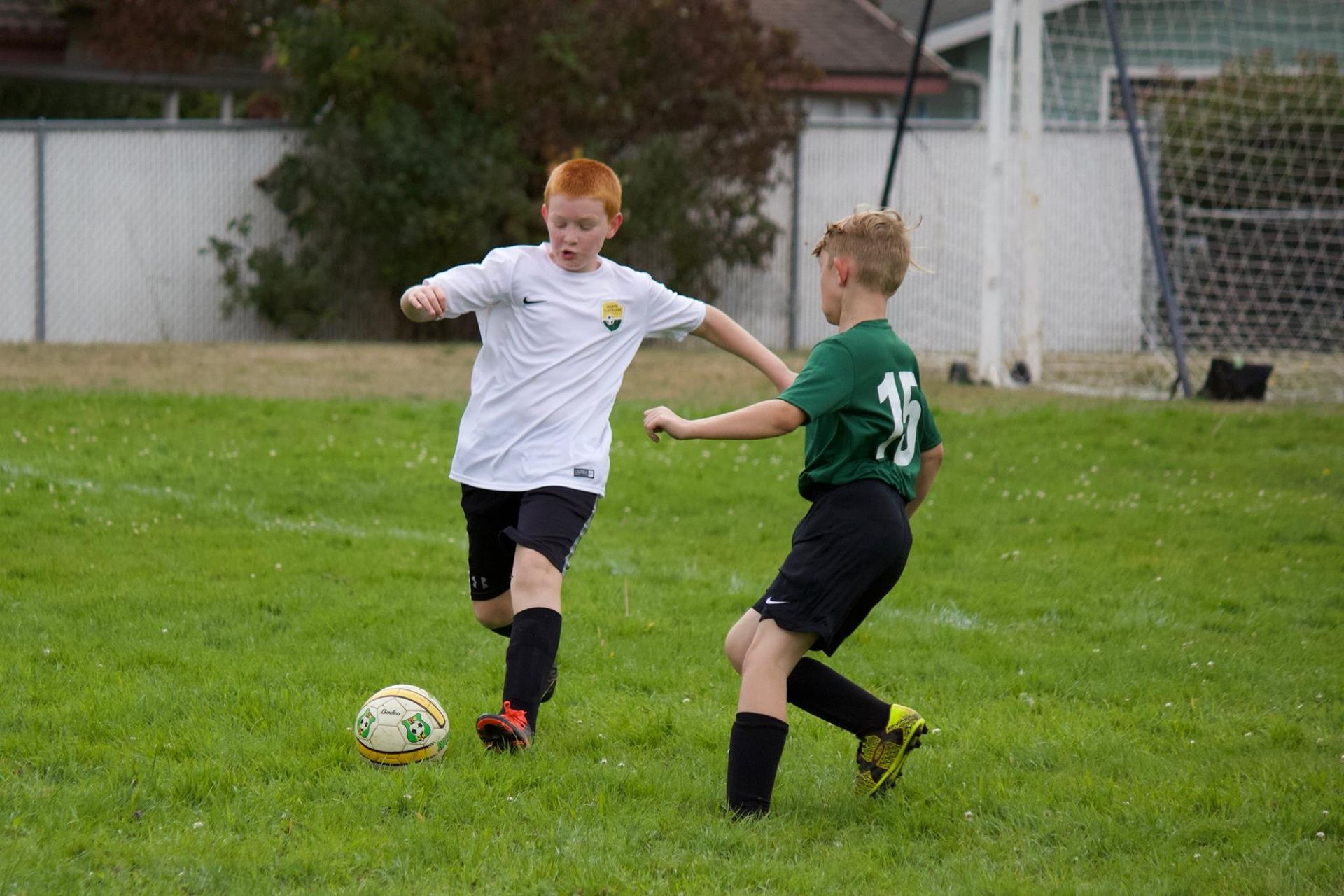Two boys playing soccer on a green field. One in white shirt dribbles ball; the other in green attempts to intercept.