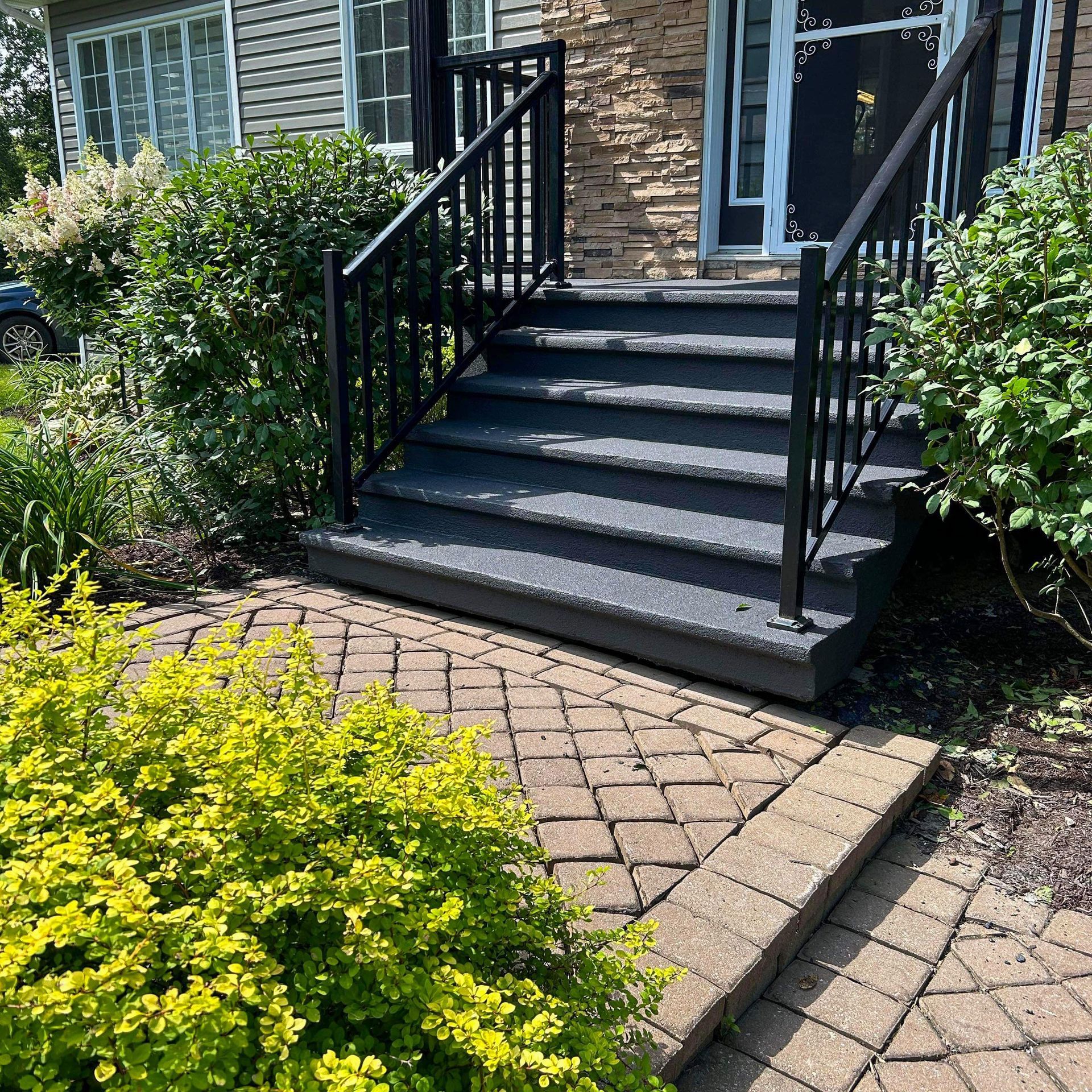 Des marches noires mènent à l'entrée d'une maison, flanquée de balustrades noires et de verdure, avec un chemin en briques.