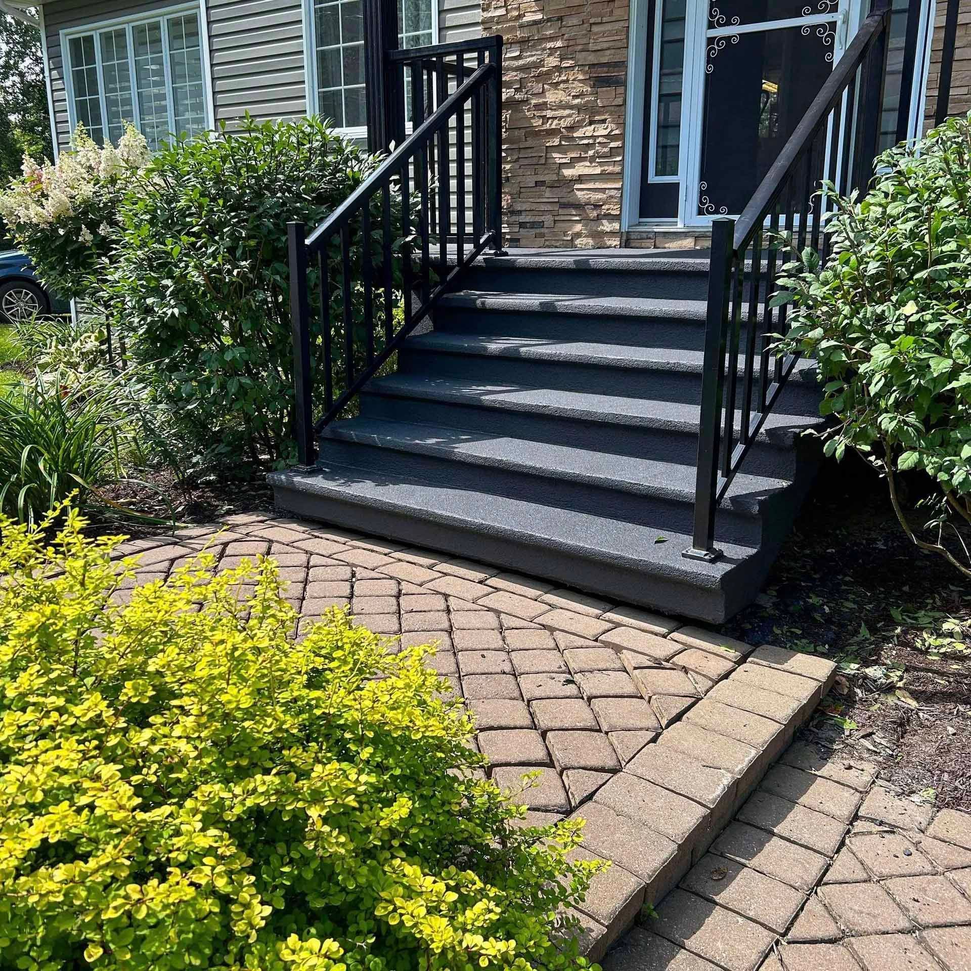 Vue extérieure des marches d'entrée d'une maison avec des rampes noires, menant à une porte d'entrée ; un chemin en briques se trouve devant.