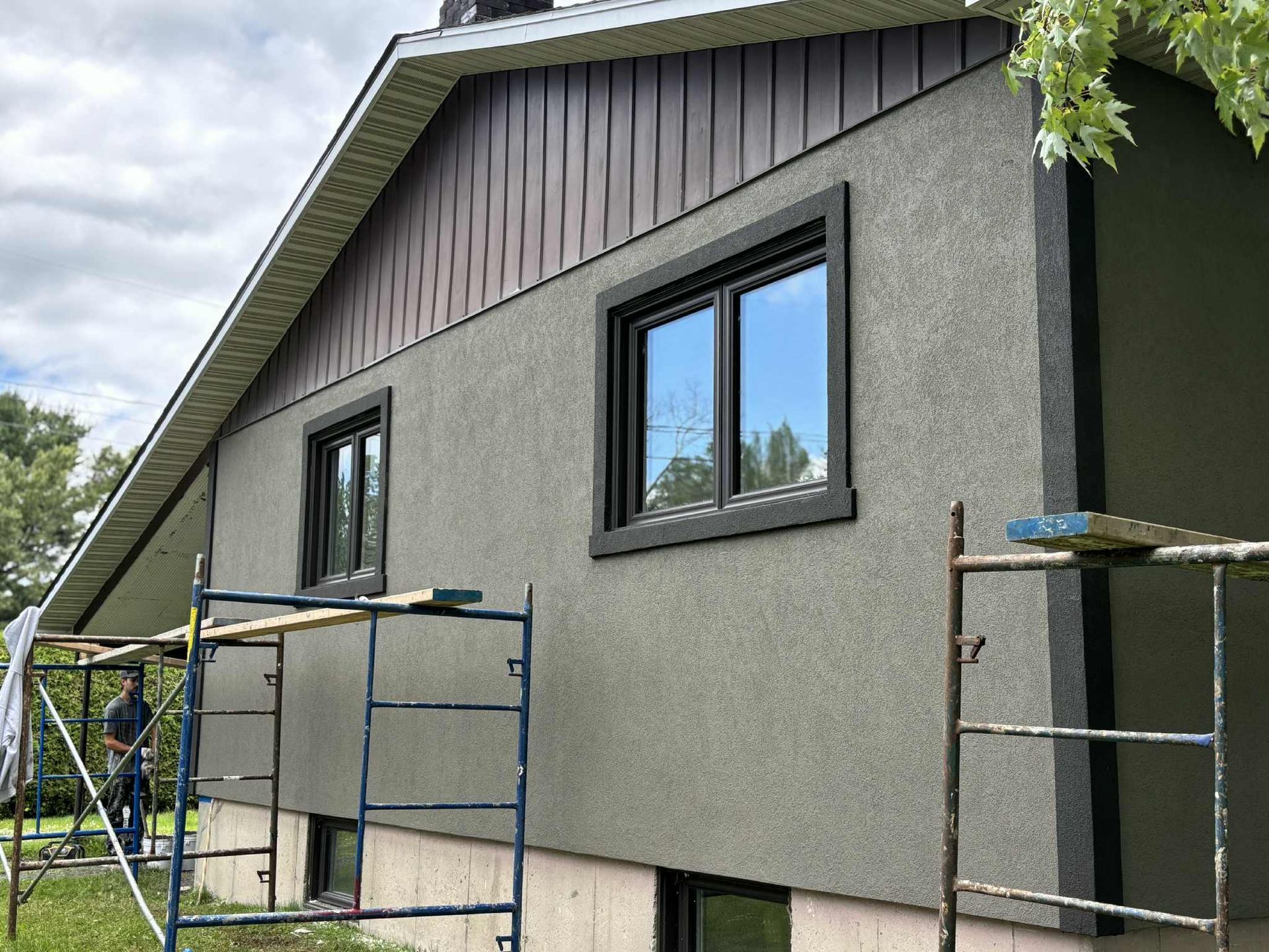Extérieur de la maison avec stuc gris, garnitures brunes, fenêtres noires et échafaudages.