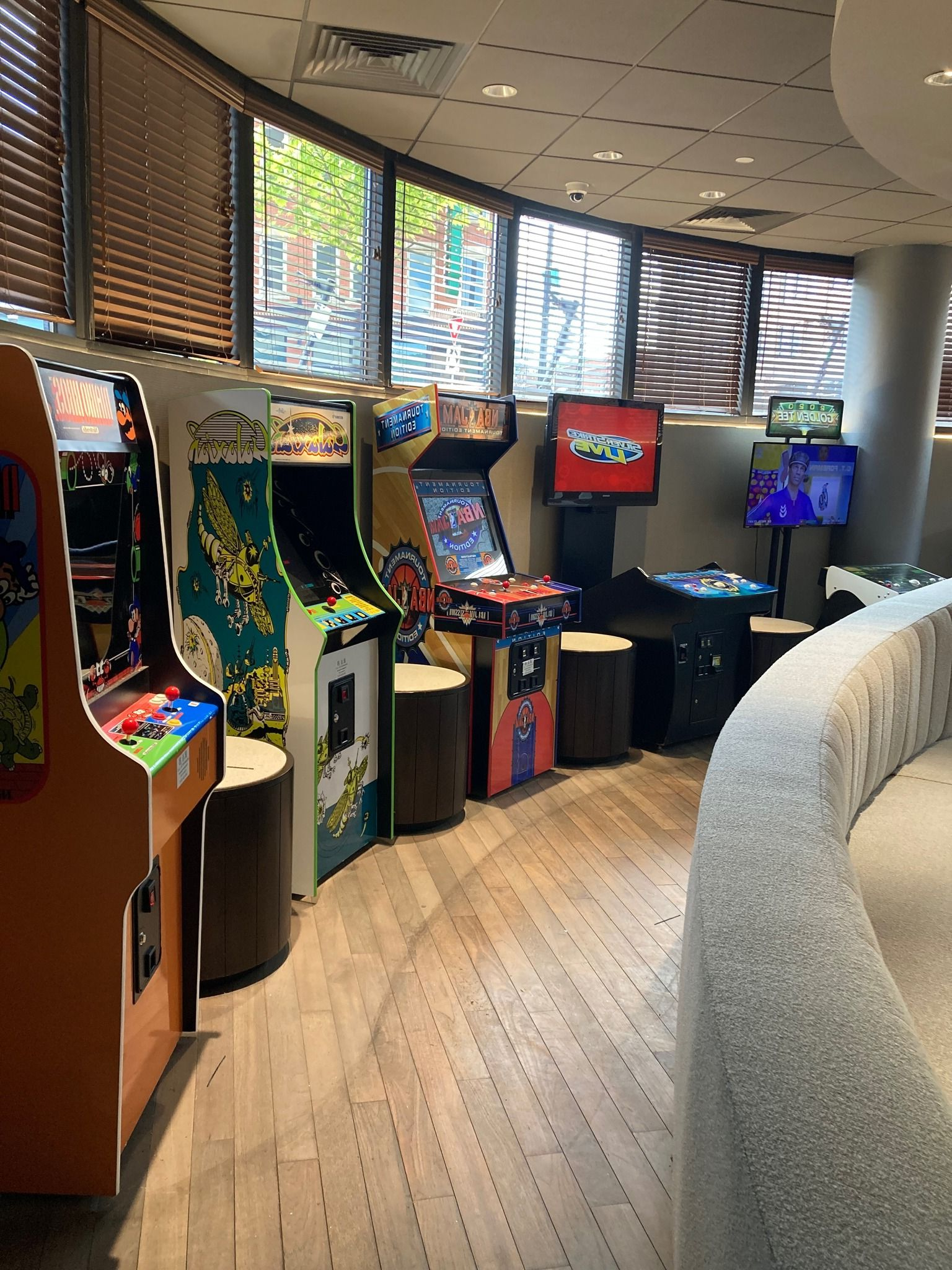 A row of classic arcade game cabinets lined up against a curved wall of large, slatted windows in a brightly lit room.