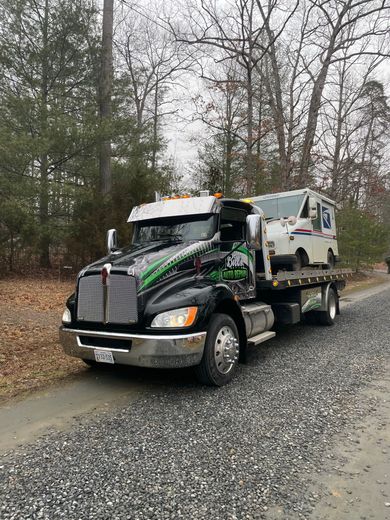 A tow truck is carrying a white van down a gravel road.