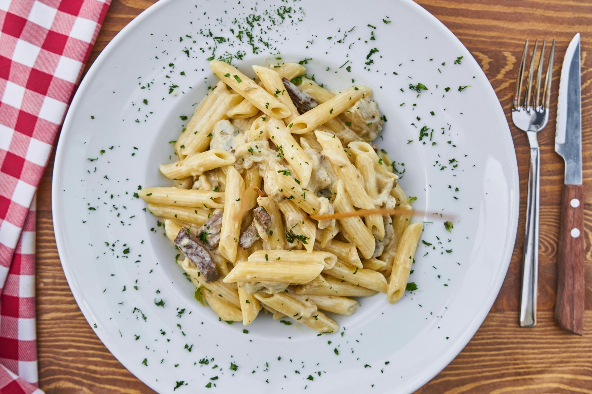 Penne pasta with sauce on a white plate, garnished with parsley, next to a knife and fork.