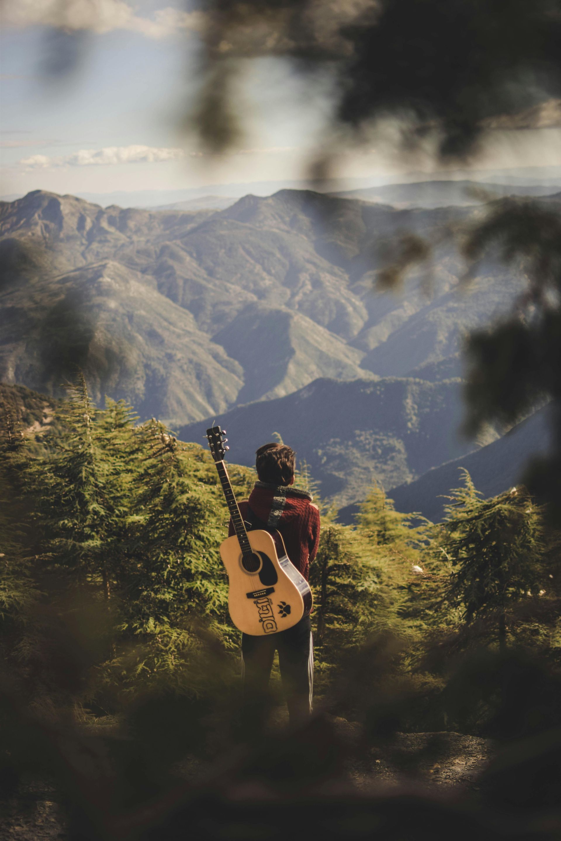 Person with guitar stands on a hilltop, overlooking mountains. The sky is bright, with green trees.