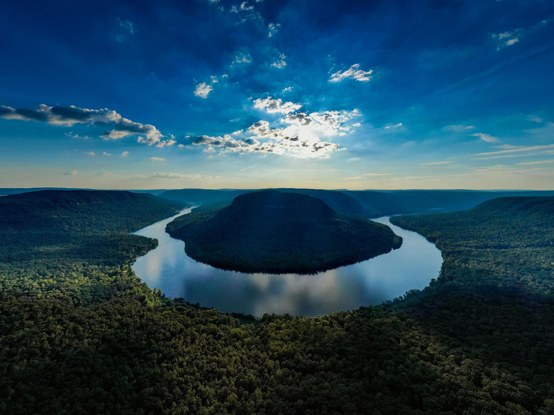 River meanders through lush green forest under a partially cloudy blue sky, sun peeking through.