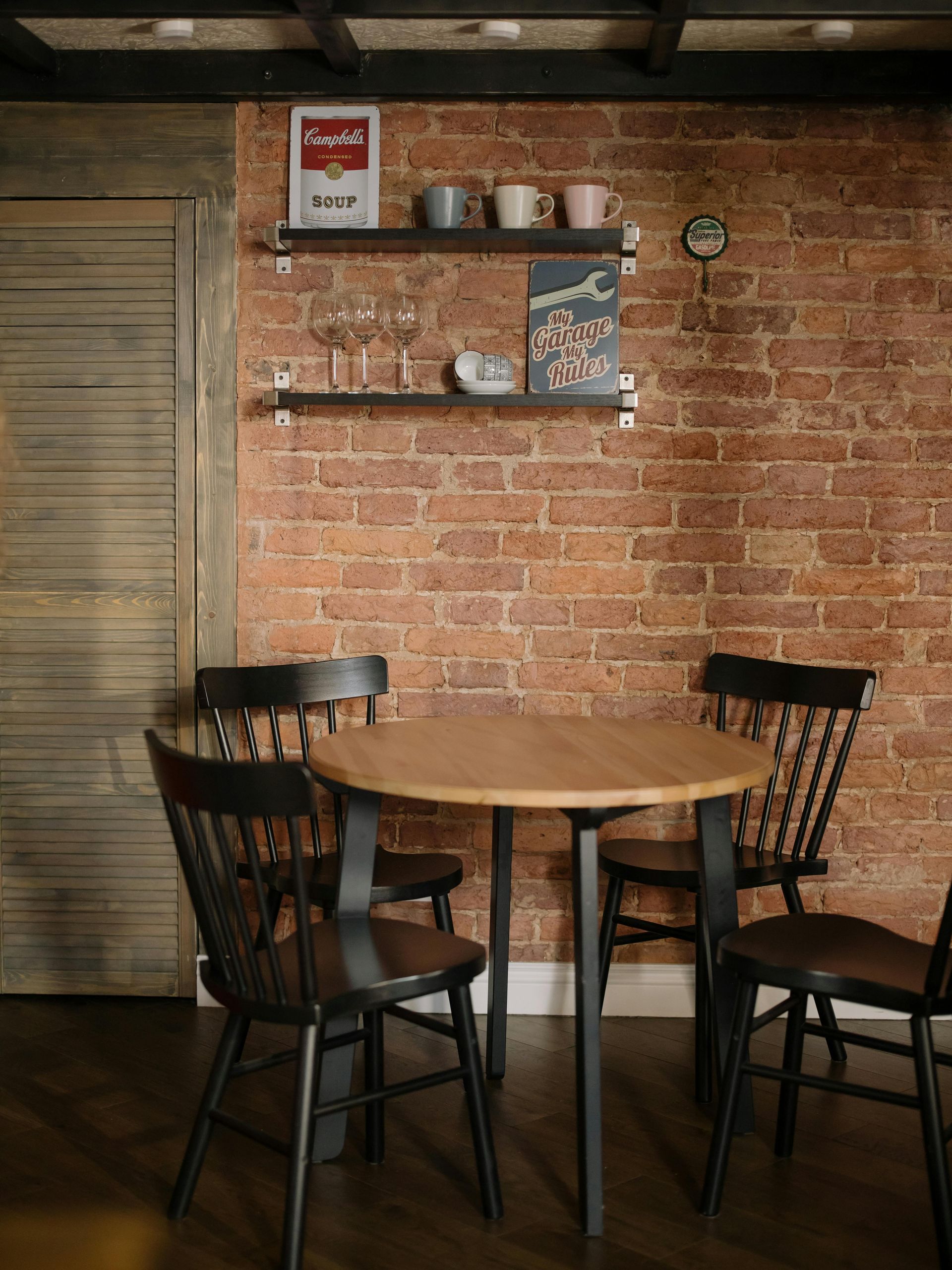 A small wooden dining table with four black chairs against a brick wall with shelves.