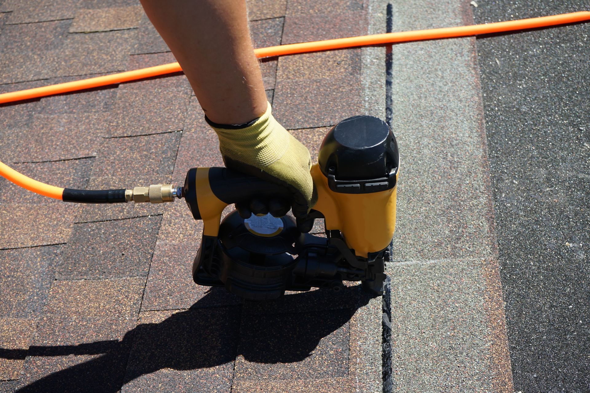 A hand uses a nail gun to nail down a roofing shingle.