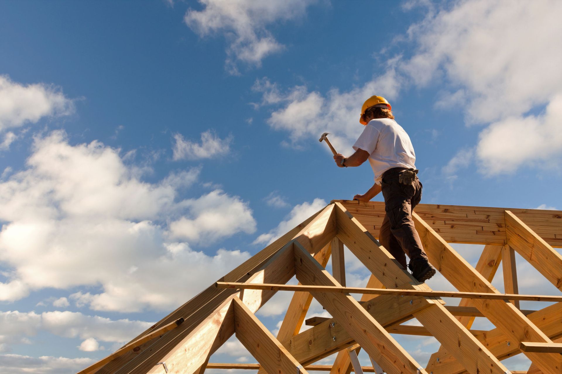 Construction worker building wooden roof frame on house under blue sky with scattered clouds.