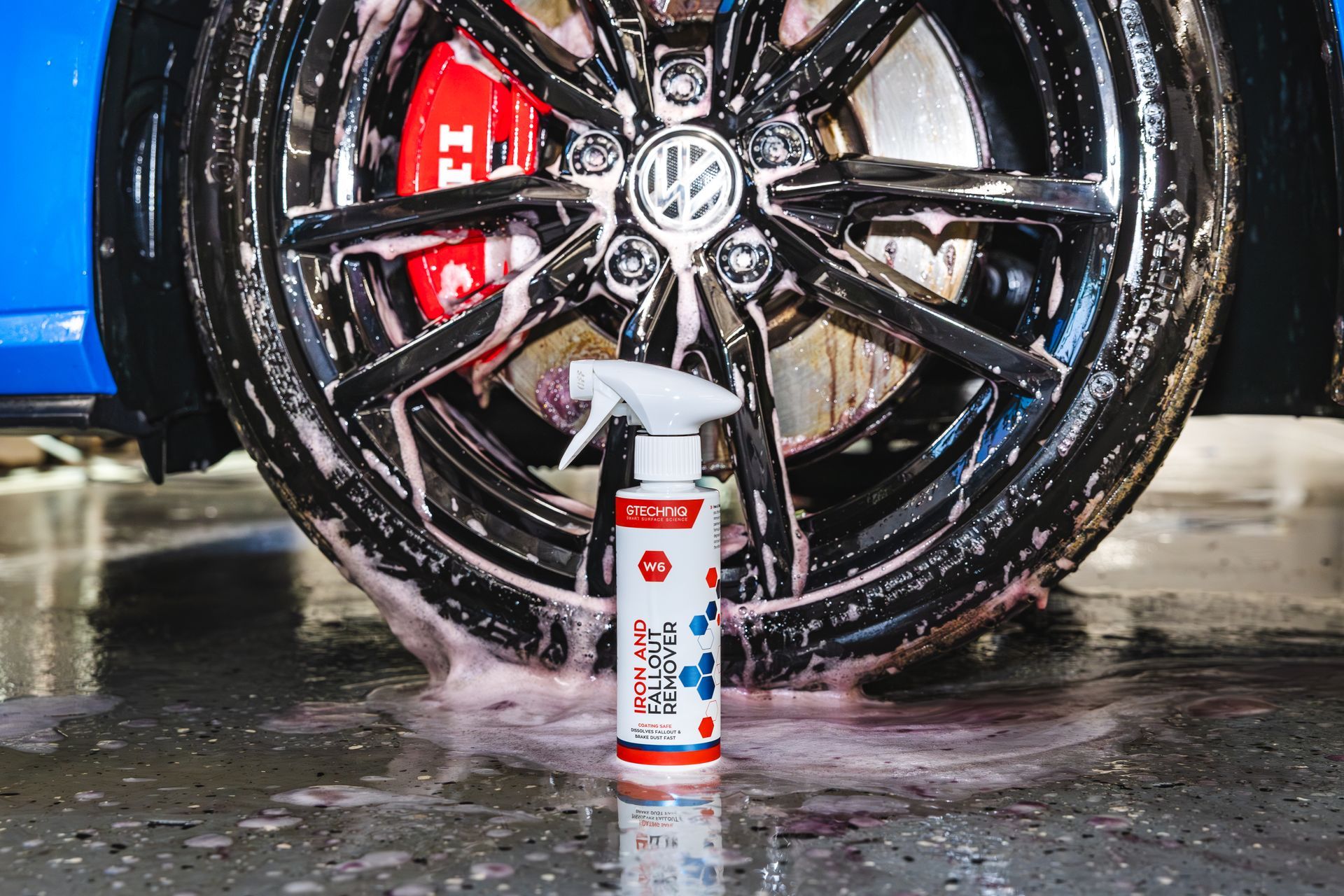 A bottle of car wash solution sits in front of a black wheel being cleaned with foamy soap.