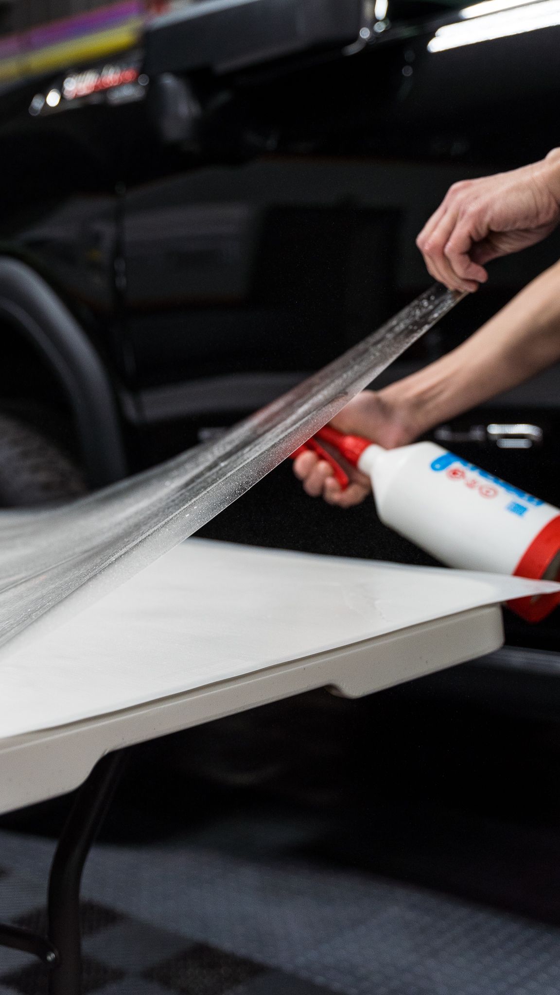 Hand peeling protective film from a white car panel with a spray bottle nearby