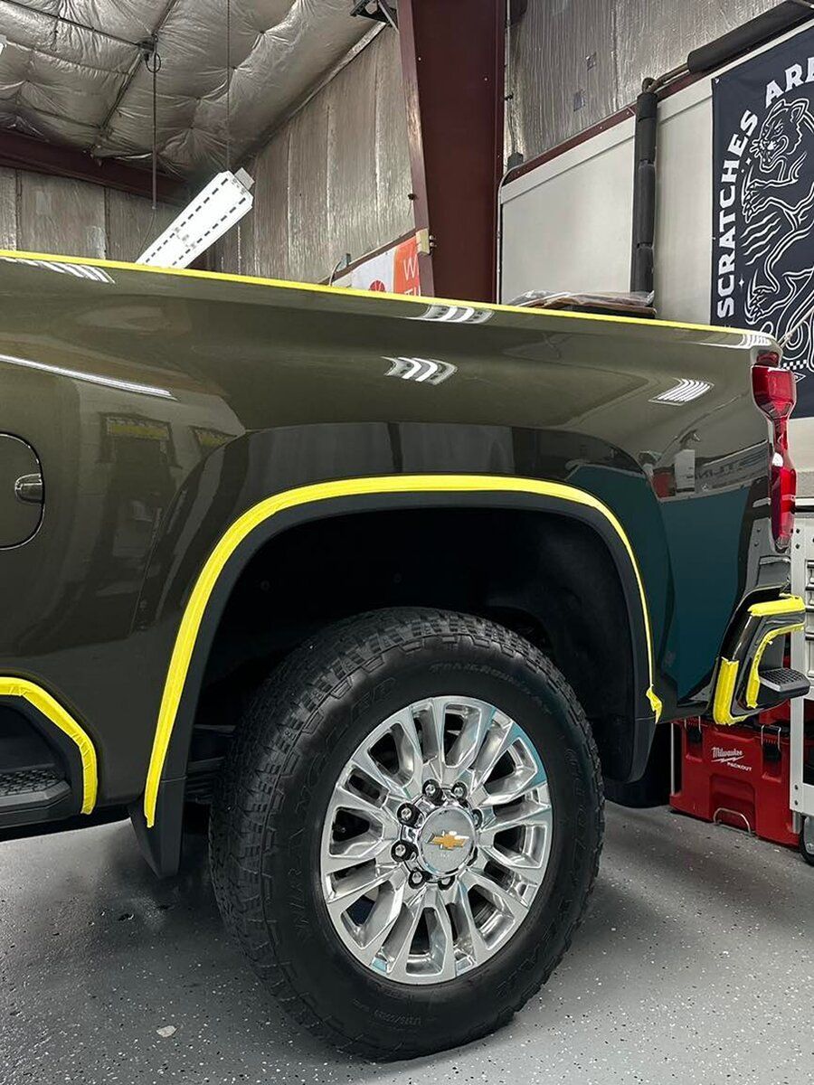 A close-up of a green Chevrolet truck with yellow tape outlining body panels. The truck is in a garage.