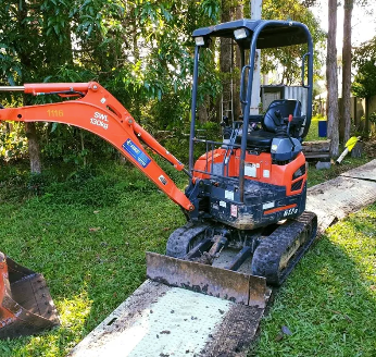 Orange mini excavator with a front bucket parked on grass beside a wooded area