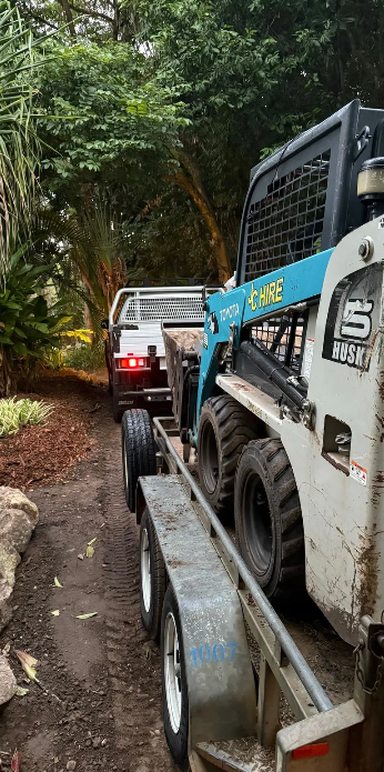Two construction vehicles on a muddy forest path, one towing the other uphill.