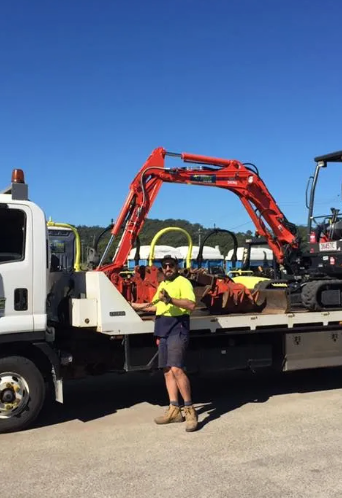 Worker standing by flatbed truck carrying red crane against blue sky