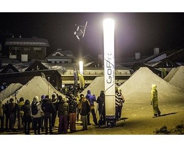 Snowboarder performing a jump at night, lit by a GoPro branded tower, surrounded by a crowd and snow piles.
