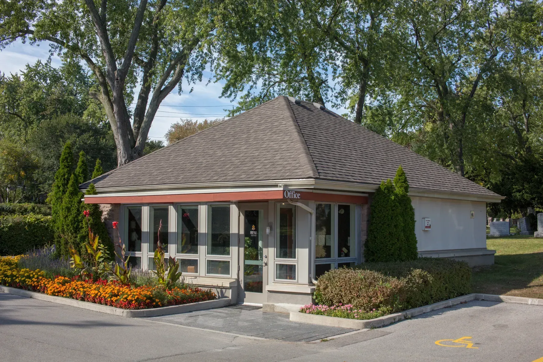Small single-story office building with a gray roof, glass entrance, and flower beds surrounded by trees