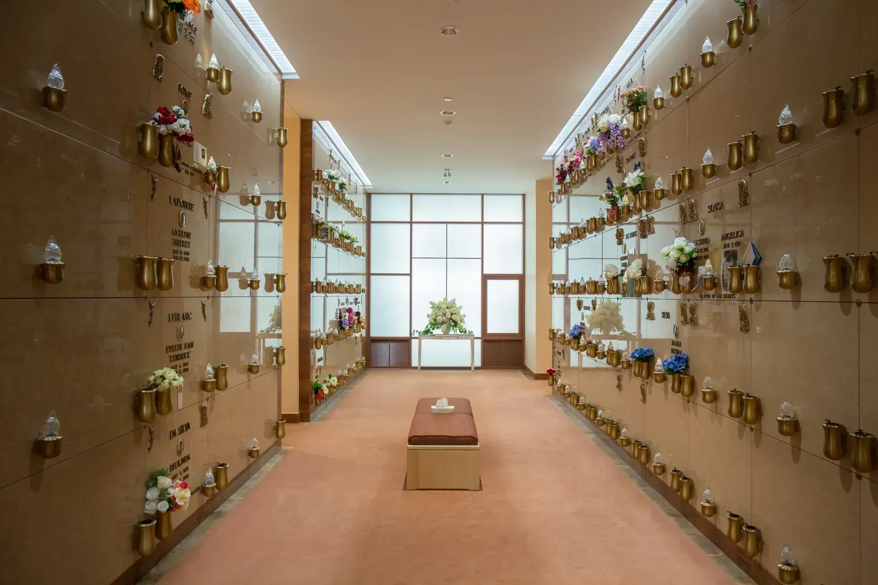 A quiet memorial columbarium corridor with rows of urn niches and flowers, lit by soft daylight.