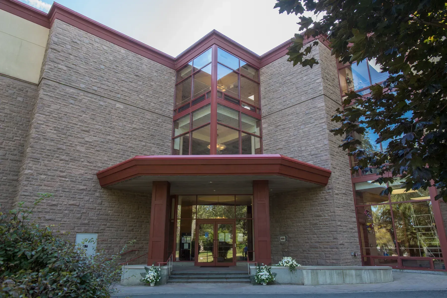 Brick building entrance with red trim and glass windows, framed by landscaping