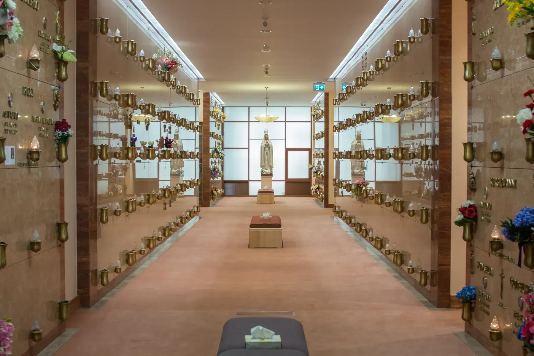 A memorial columbarium corridor with rows of niches and flowers leading to a statue at the far end.