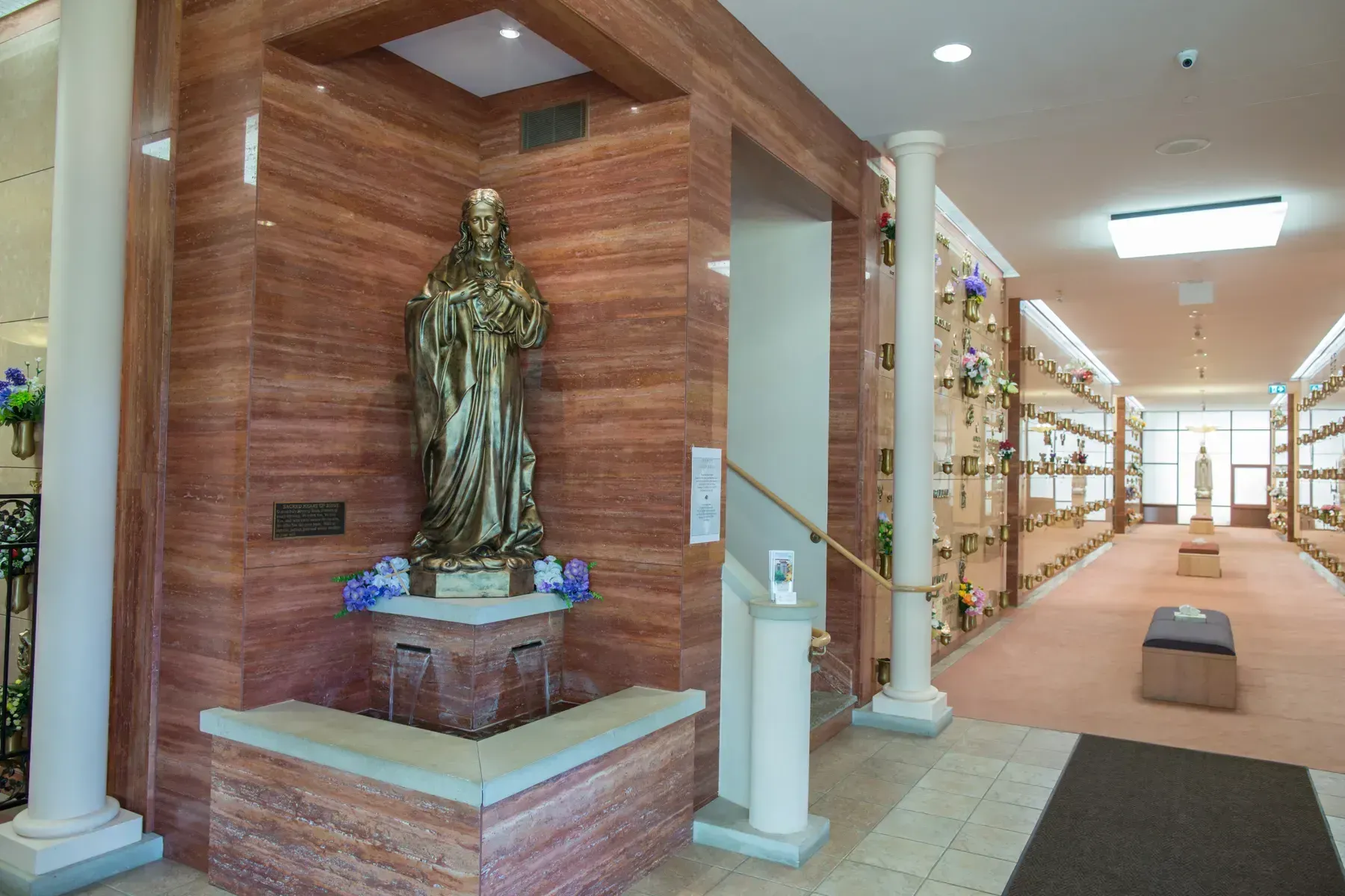Bright chapel corridor with a statue in a lit niche, columns, and rows of candles along the wall