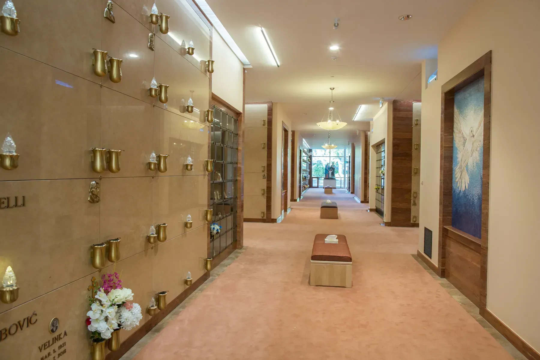 Long indoor corridor with wall-mounted brass plaques and framed art, carpeted floor, and benches.