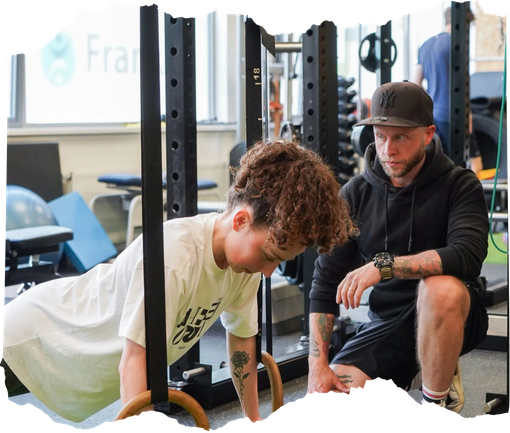 Een trainer begeleidt iemand bij het doen van push-ups in een sportschool, omringd door fitnessapparatuur.