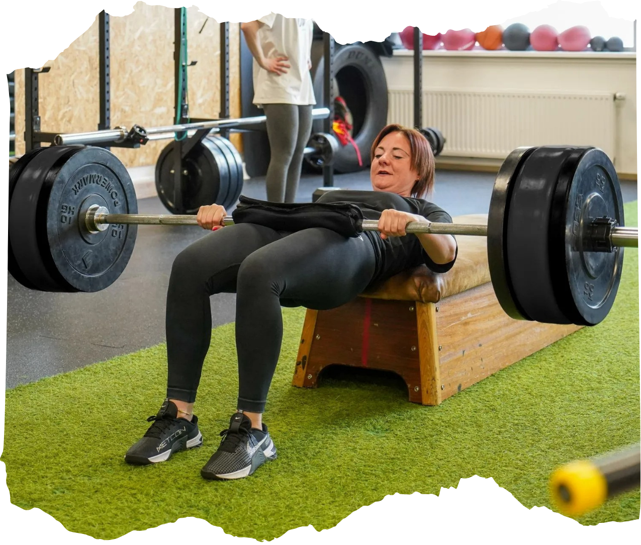 Vrouw voert heupstoten uit met een halter in een sportschool, liggend op een bankje op kunstgras.