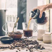 Coffee being poured into a clear mug beside coffee beans and brewing tools on a bright kitchen counter
