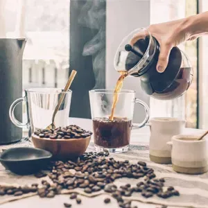 Coffee being poured into a clear mug beside coffee beans and brewing tools on a bright kitchen counter