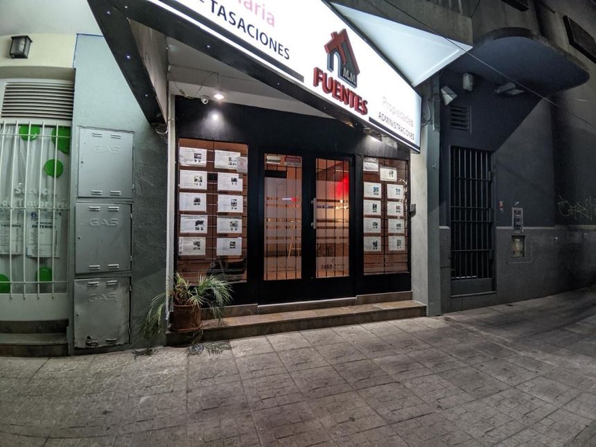 Fachada de tienda con letreros iluminados y entrada de cristal en una acera de la ciudad por la noche.