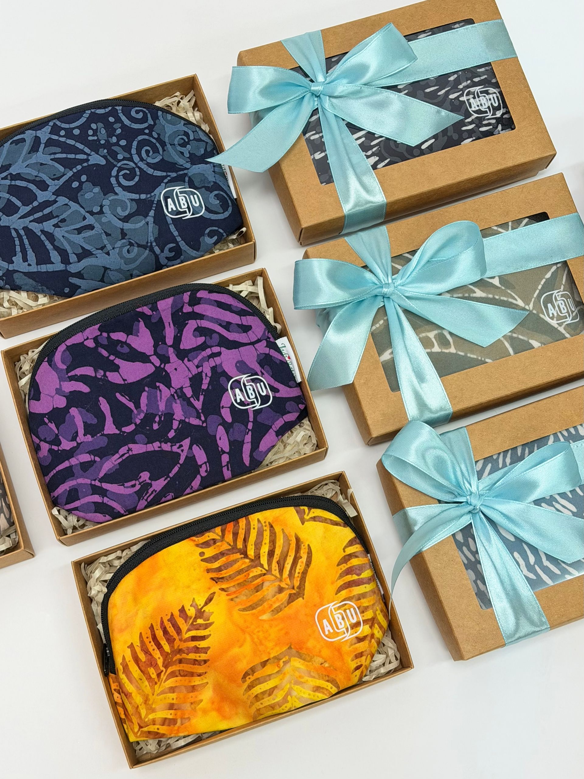 Handmade Malaysian batik fabric pouches in assorted colours displayed in kraft gift boxes with blue ribbon packaging, arranged as elegant batik gift sets for corporate gifting, events, and special occasions.