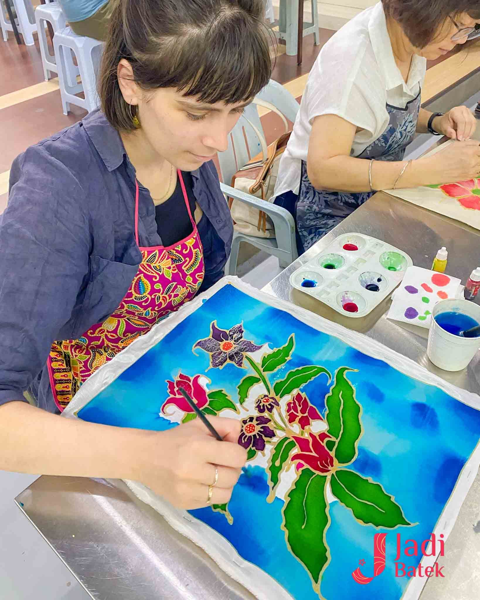 Woman painting batik art with colorful flowers and a teal background.