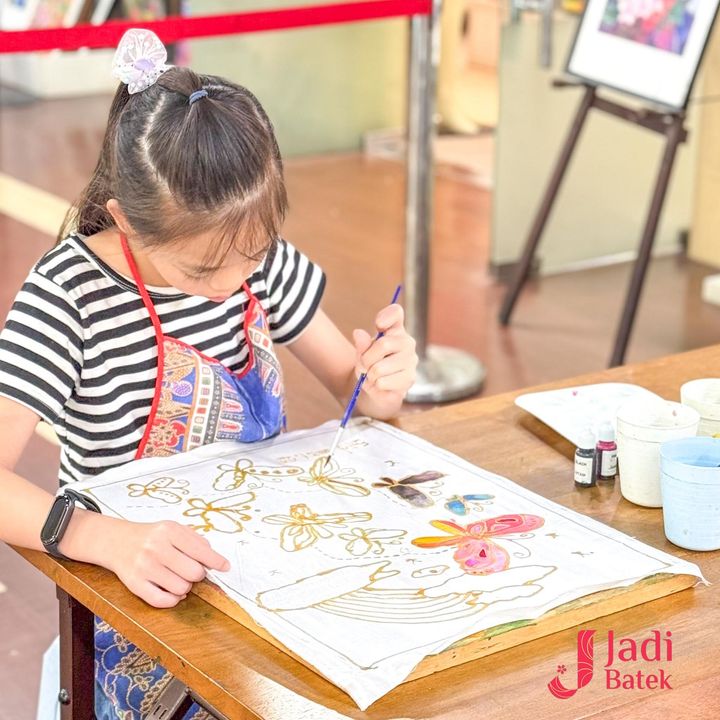 Girl paints batik with brush at wooden table. Wearing striped shirt, apron, and hair bow.