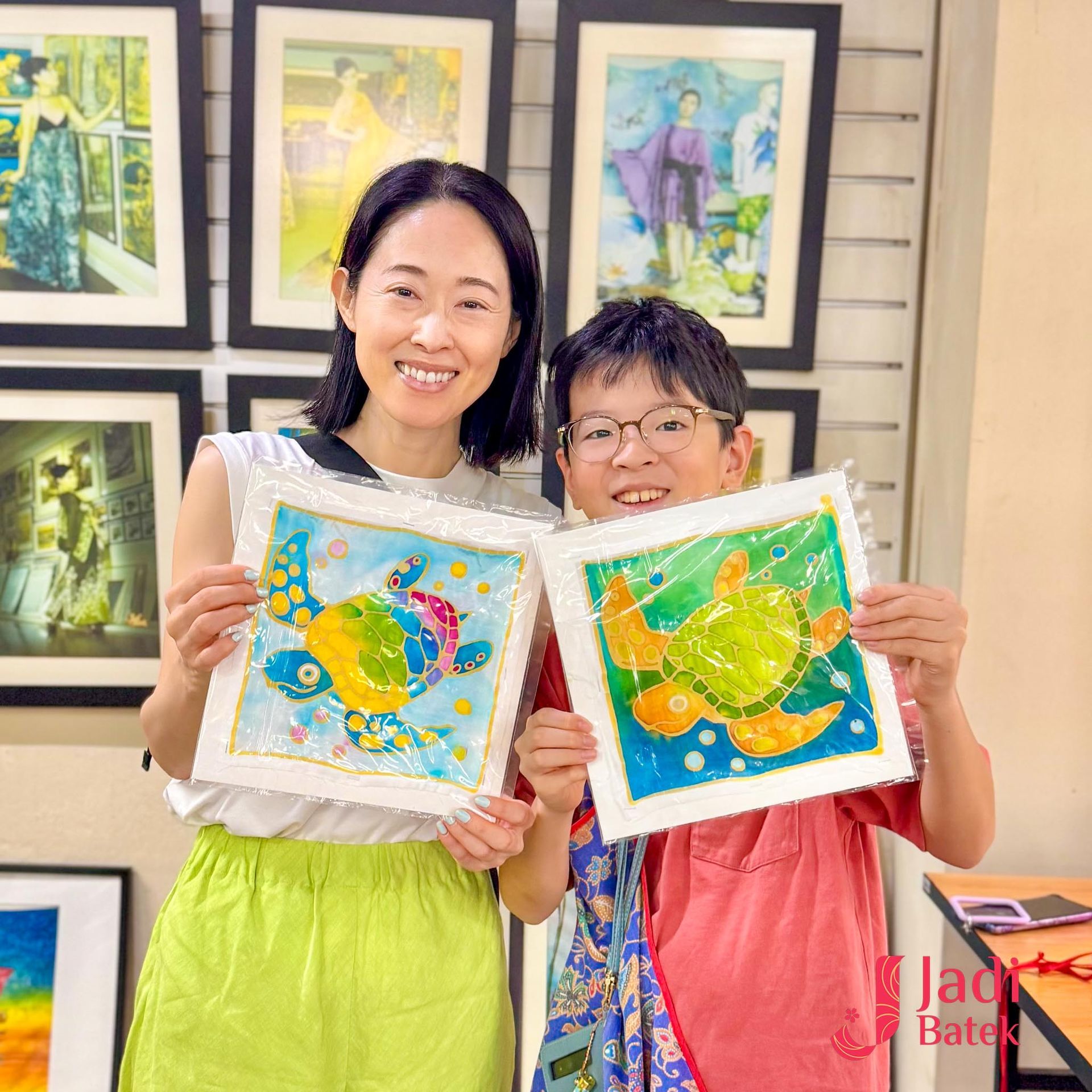 Woman and boy smiling, holding up colorful turtle paintings in art studio.
