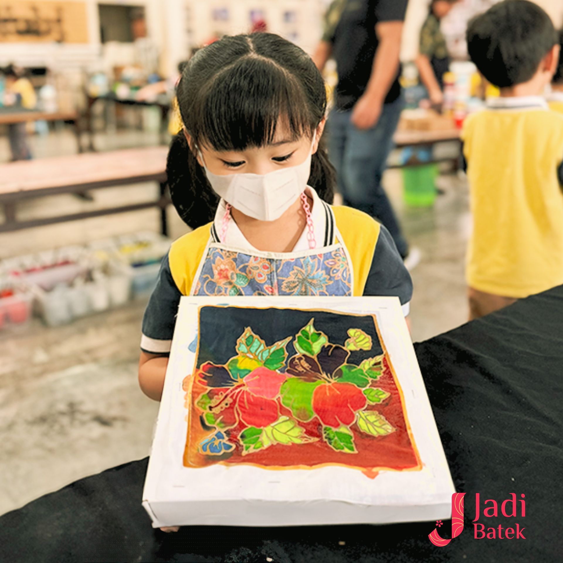 Girl in mask holding a batik painting of flowers; indoor art workshop.