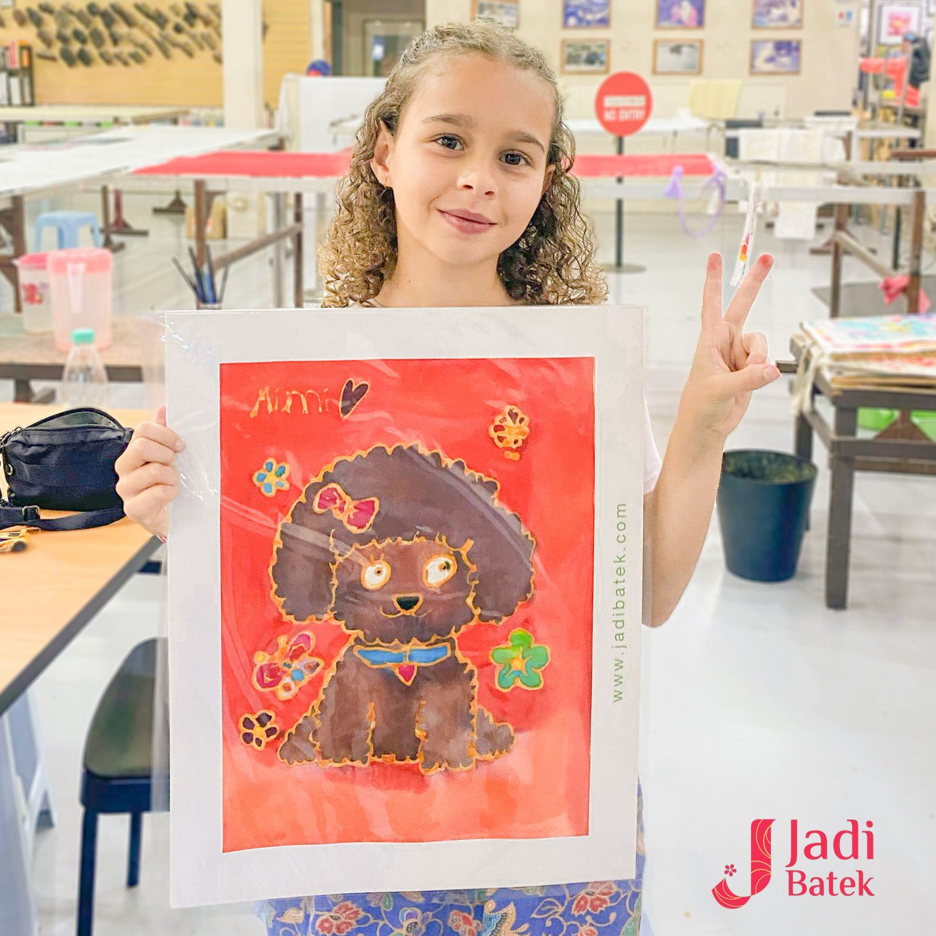 Young girl holding a framed dog painting, smiling and giving a peace sign. Batik workshop setting.