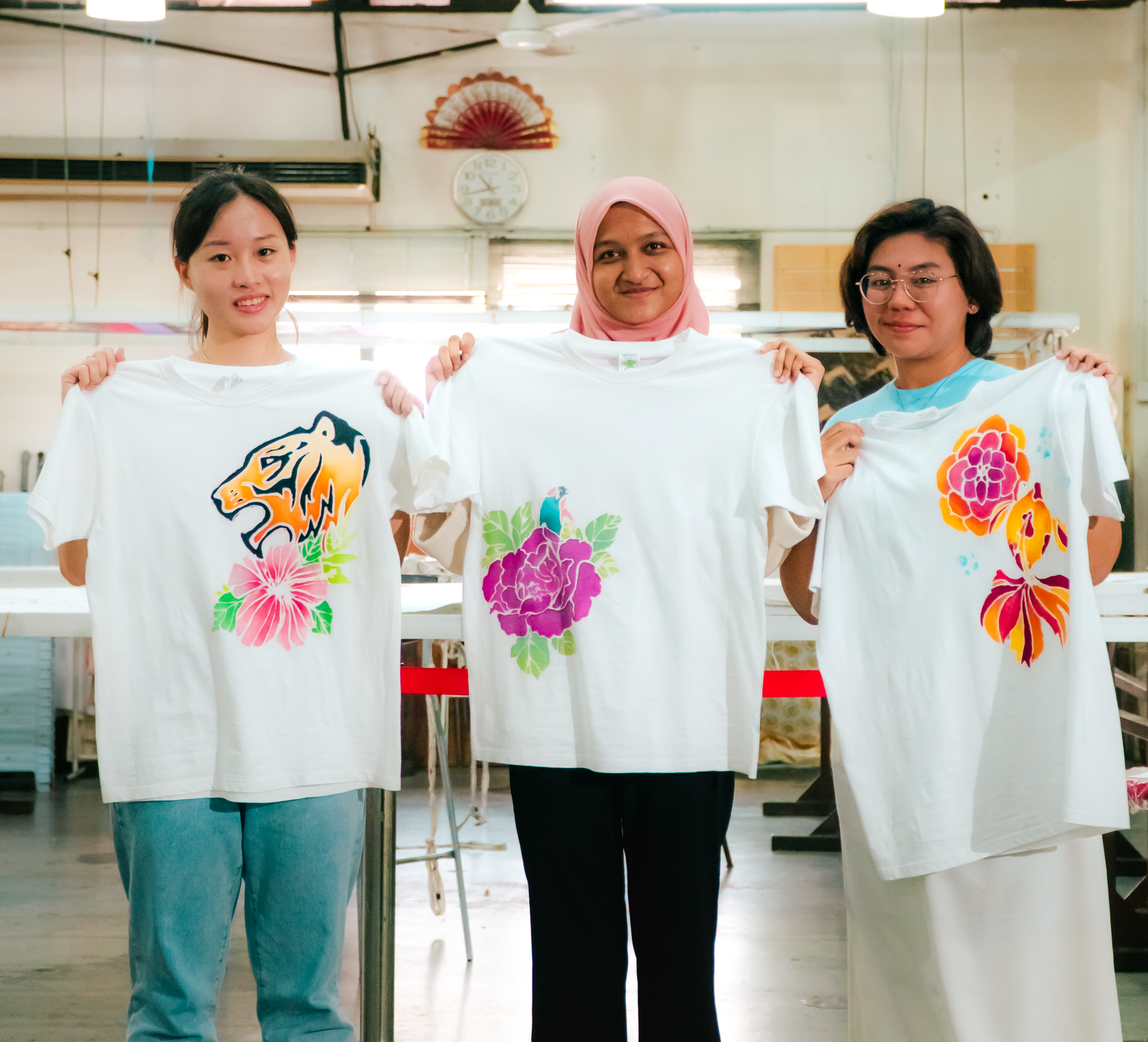 Three women holding up custom-painted white t-shirts with floral and animal designs.