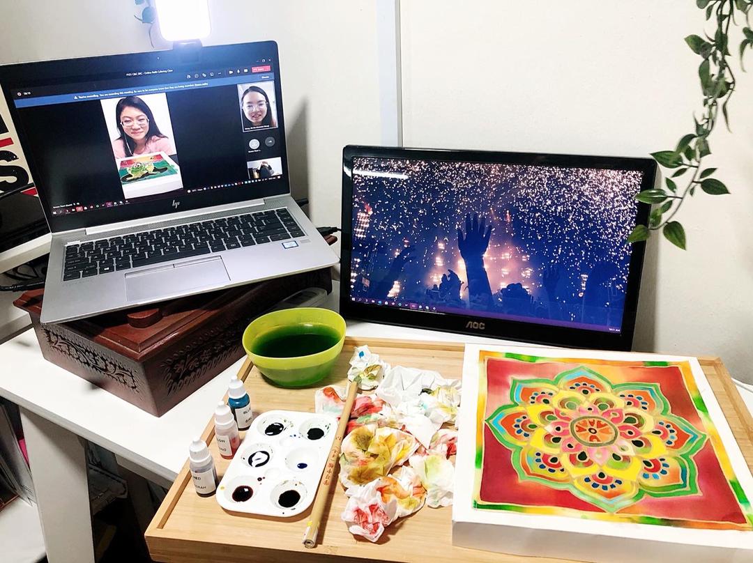 A person painting, with a video call on laptop, paint supplies, and a finished artwork on a white table.