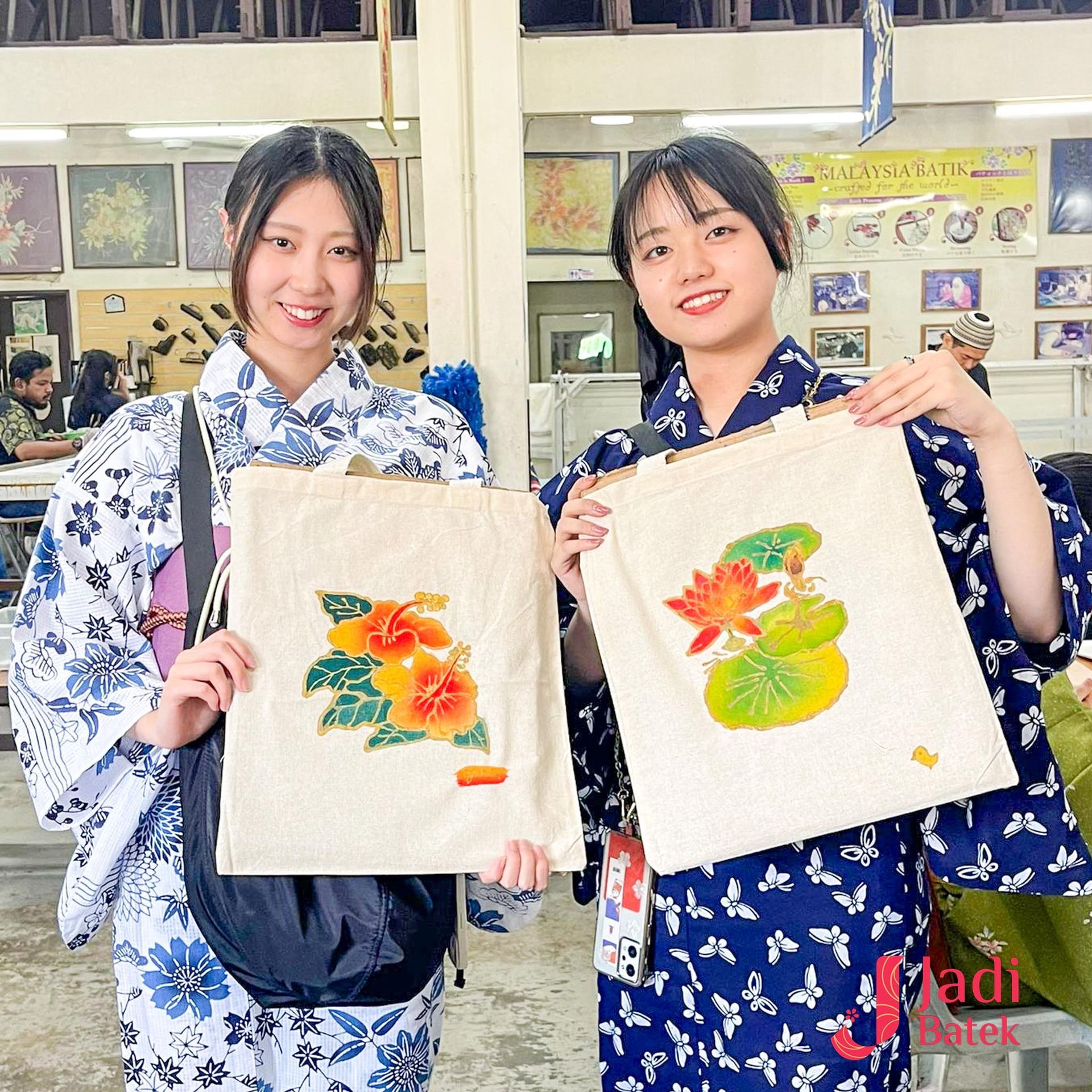 Two women in yukatas display tote bags with painted flowers. They smile in a workshop setting.