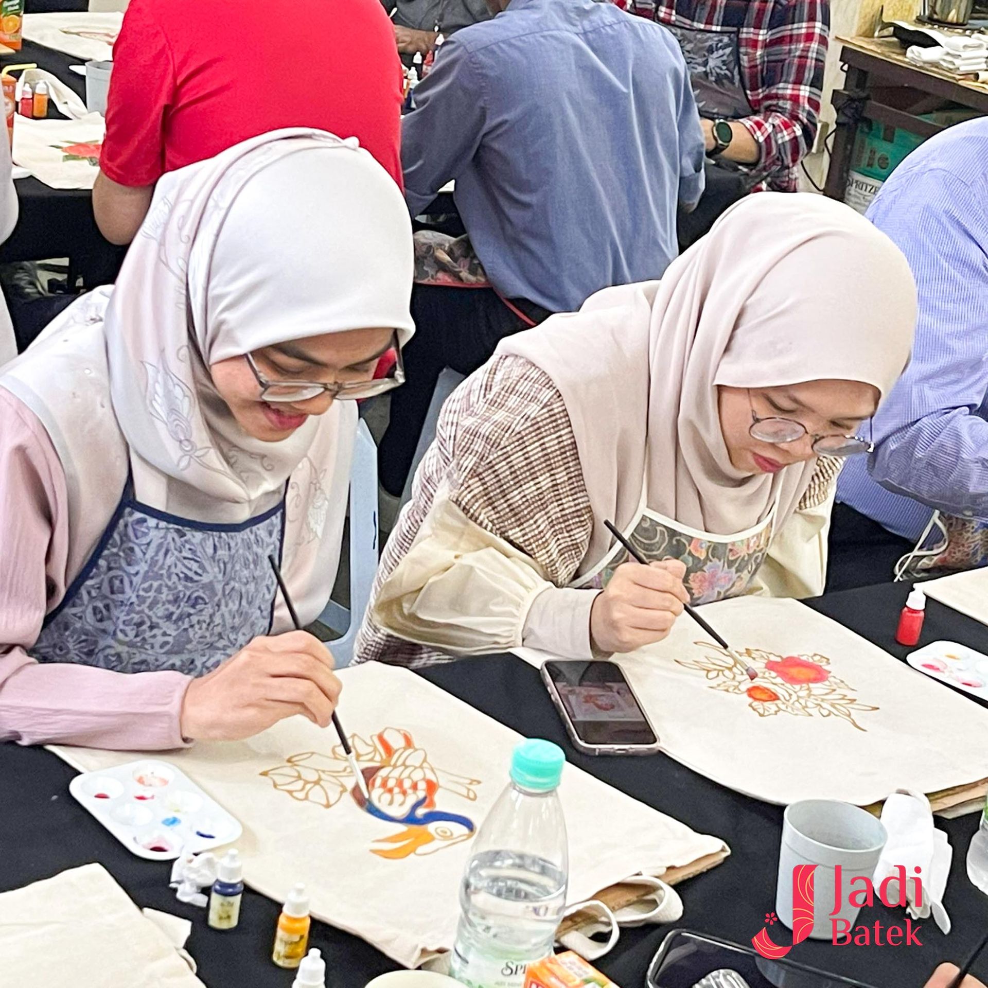 Two women in headscarves paint on fabric, smiling, at a workshop.