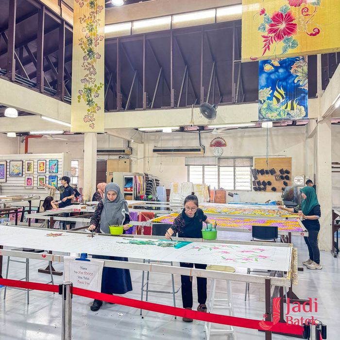 People painting batik fabric in a workshop; white tables, colorful textiles hang above.
