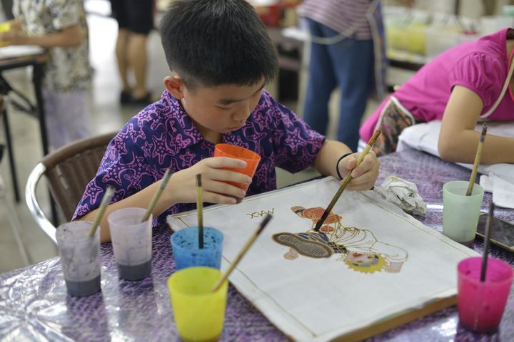 Boy painting with brush, dipping it in a cup, focused on art project with colorful cups in front of him.